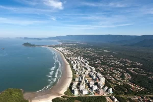 Vista aérea da Praia da Riviera, em São Lourenço, mostrando a faixa de areia, prédios à beira-mar e áreas verdes ao fundo.