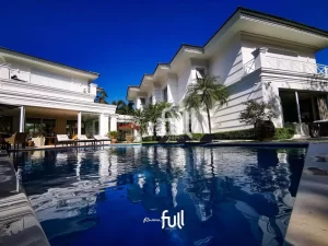 Mansão branca de dois andares com piscina ampla em primeiro plano, rodeada por espreguiçadeiras, palmeiras e móveis de área externa sob céu azul claro.