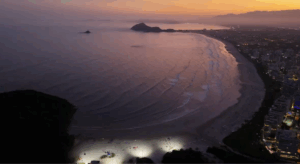 Vista aérea da Riviera de São Lourenço ao entardecer, mostrando a orla iluminada, o mar com pequenas ondas e a cidade ao fundo.