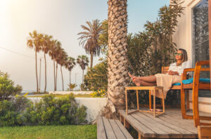 Mulher de óculos de sol relaxando descalça em cadeira de madeira em deck ensolarado, com vista para o mar e coqueiros
