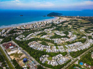 Vista aérea de uma cidade litorânea com prédios à beira-mar, bairros planejados com casas e condomínios rodeados por áreas verdes, e o oceano azul com pequenas ilhas ao fundo.