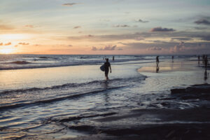 Pessoa caminhando na beira do mar ao pôr do sol, segurando uma prancha de surfe.