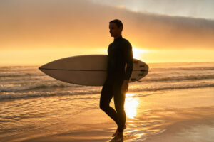 Pessoa caminhando na praia ao pôr do sol com prancha de surf, em silhueta, com o mar e a luz dourada ao fundo.