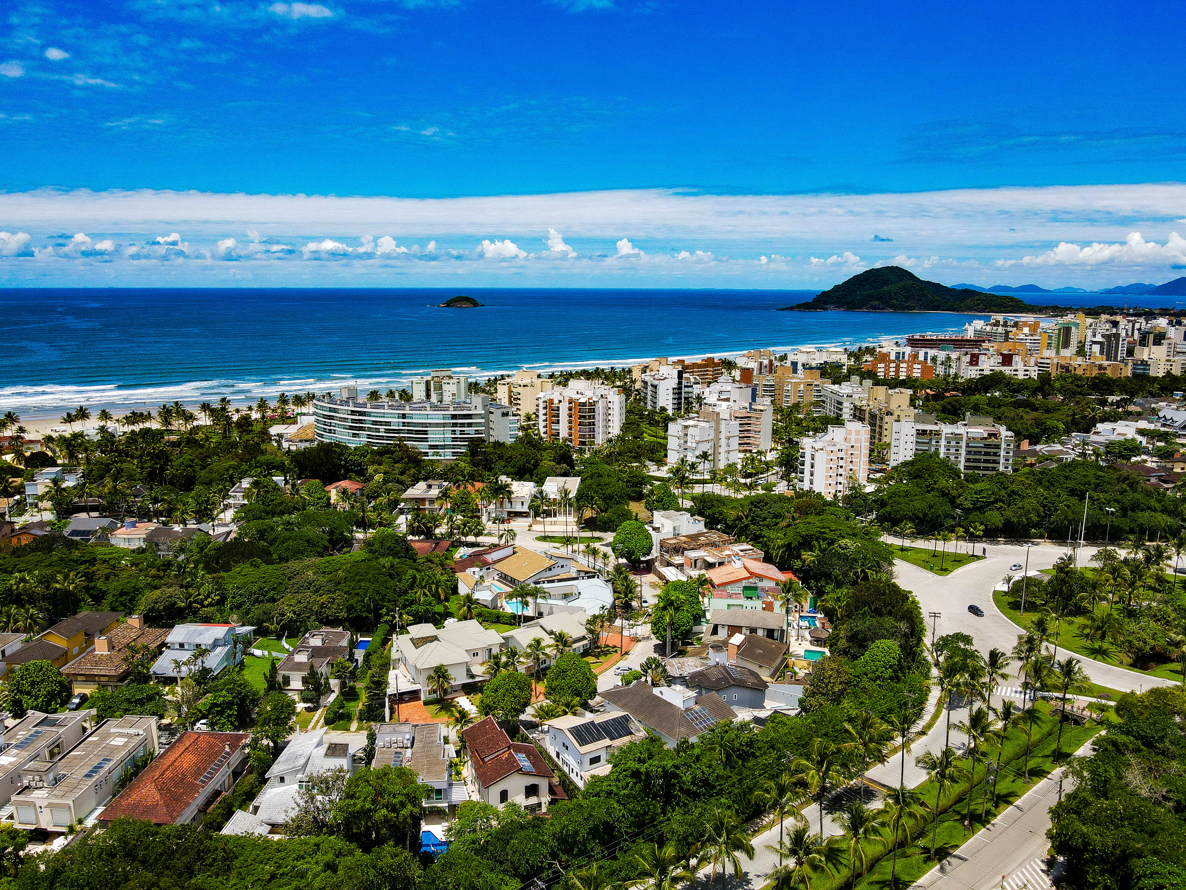Imagem aérea de uma cidade litorânea com prédios, casas, muitas áreas verdes e a praia ao fundo, com mar azul e céu claro.