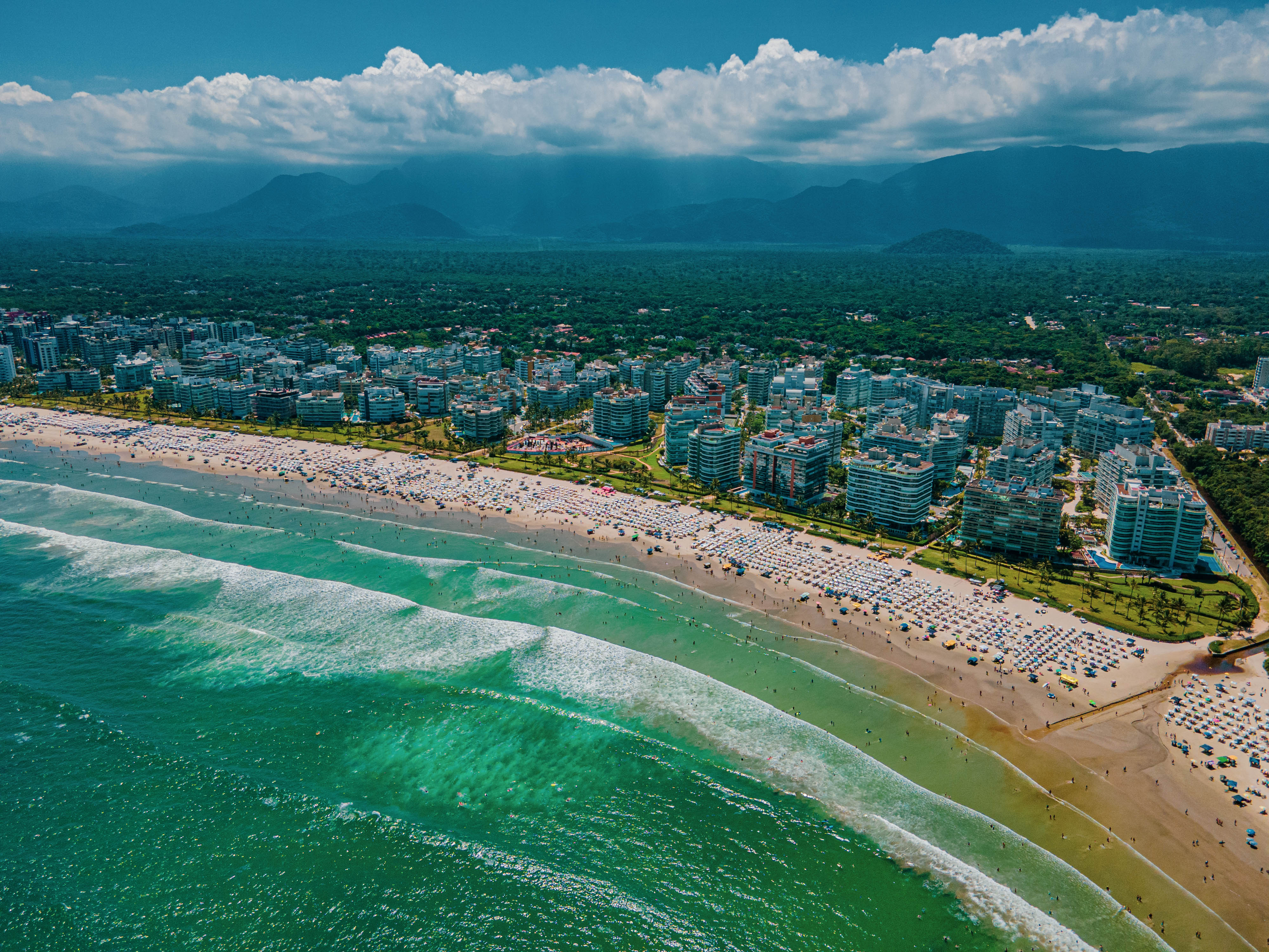 Imagem aérea da Riviera de São Lourenço, mostrando a faixa de areia organizada, o mar, prédios residenciais alinhados à orla e extensa área de vegetação com a Serra do Mar ao fundo.