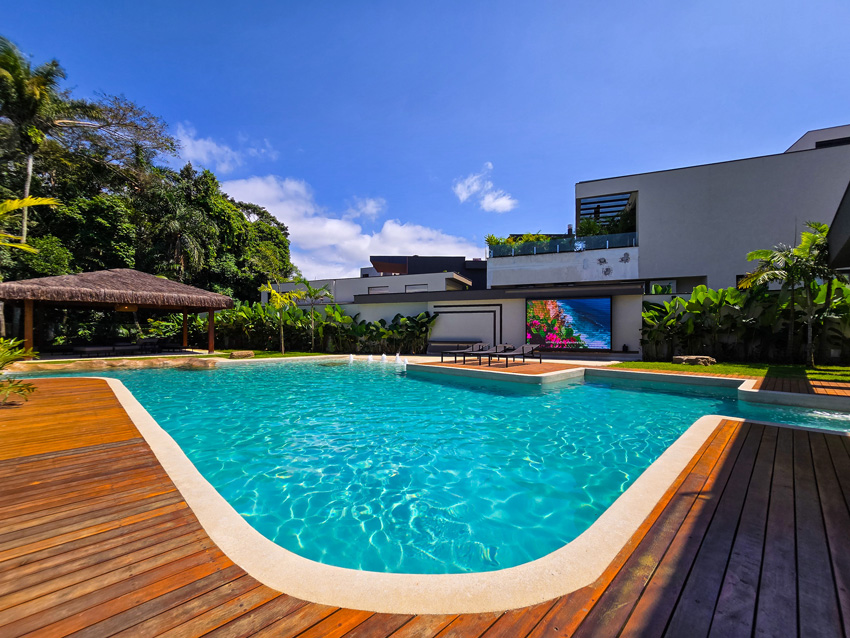 Piscina ampla com deck de madeira em área externa de residência moderna, cercada por jardim tropical e céu azul.