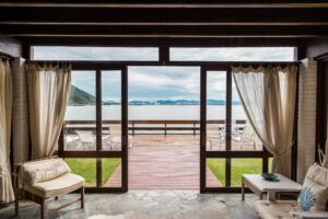 Sala com portas de vidro abertas para um deck de madeira à beira de um lago, com cadeiras brancas ao ar livre e vista para montanhas ao fundo.