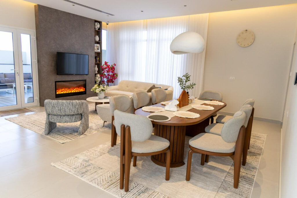 Sala de estar e jantar integrada em estilo moderno e minimalista. O ambiente possui tons neutros, com uma mesa de jantar de madeira oval ao centro, cadeiras estofadas claras, uma lareira elétrica sob a TV e uma grande luminária pendente branca.