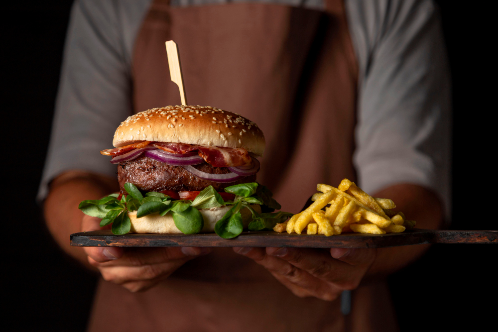 Hambúrguer artesanal gourmet com bacon, cebola roxa e folhas verdes, servido com batatas fritas em uma tábua de madeira.