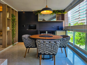 Varanda gourmet moderna com mesa redonda de madeira, poltronas estofadas, churrasqueira embutida e ampla janela com vista para prédios residenciais.