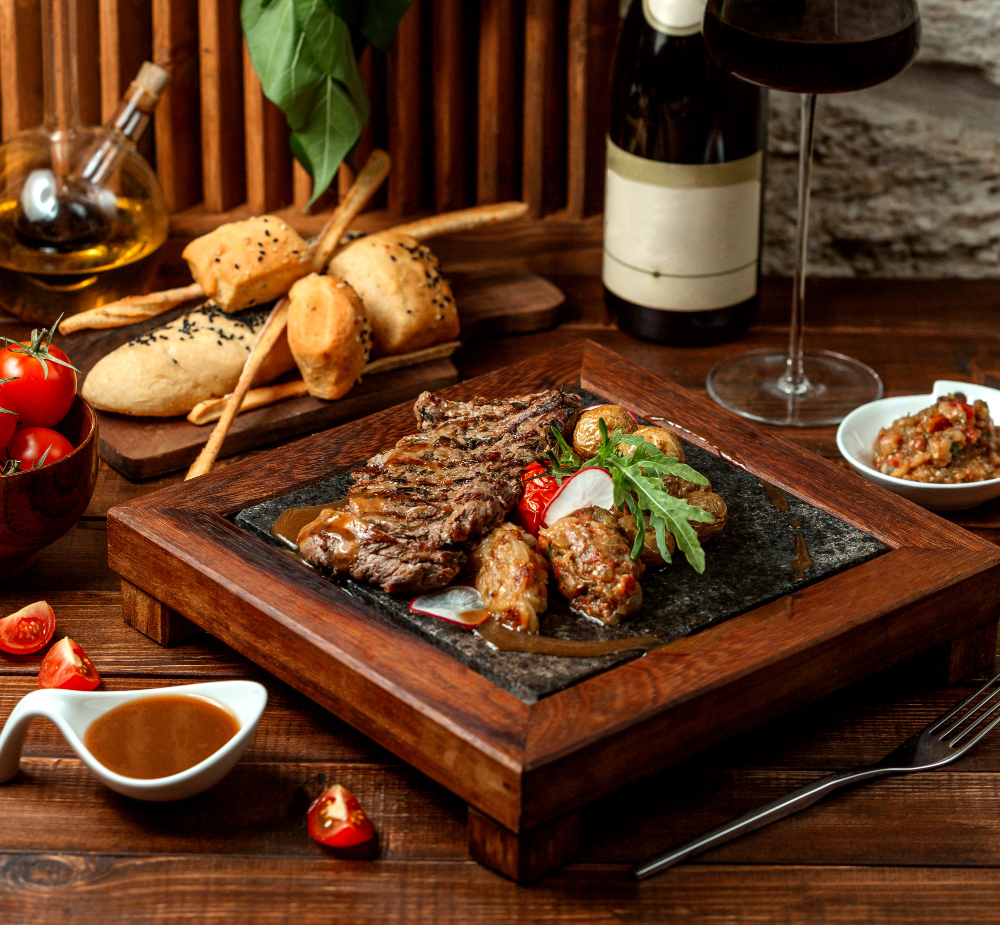 Prato de carne grelhada servido em chapa de pedra com acompanhamentos, pão artesanal e taça de vinho tinto sobre mesa de madeira.